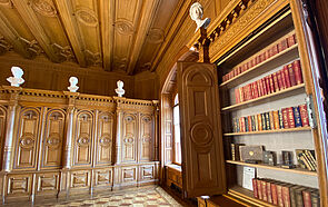 In der Bibliothek stehen Holzschränke an der Wand. Einer ist geöffnet. Darin befinden sich antike Bücher. Die Decke des Raumes besteht aus Holz.