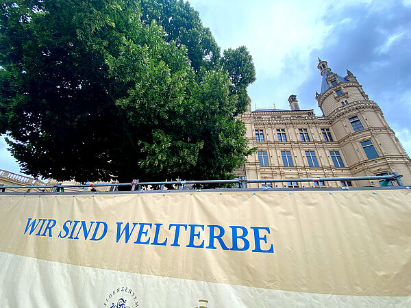 Ein Banner mit dem Titel „Wir sind Welterbe" an einem Bauzaun, hinter dem sich das Schweriner Schloss und ein Kastanienbaum erheben.