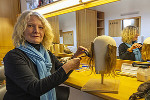 Eine Frau sitzt vor einem Spiegel und frisiert die Haare einer Perücke. 