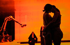 Bühnenbild im orangenen Licht. Zu erkennen sind die Silhouetten von mehreren Künstlerinnen und Künstlern.