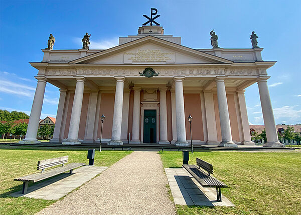 Eine Wiese. Dazwischen ist ein Weg. Links und rechts des Weges steht eine Bank. Am Ende des Weges steht die Stadtkirche. 