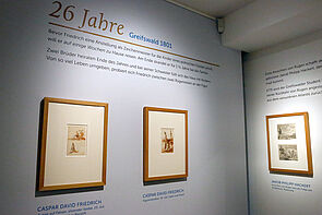 Drei Bilder im Holzrahmen hängen an zwei Wänden. Zu sehen sind insgesamt vier kleinformartige Zeichnungen.