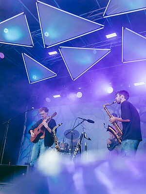 Zwei junge Männer stehen auf einer Bühne. Sie spielen Saxophon und Gitarre. Der Raum ist blau erleuchtet. 