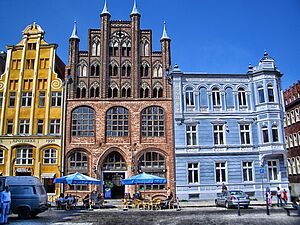 Ein gelbes und backsteinfarbenes Giebelhaus. Daneben ein blaues Eckhaus. Davor: der Marktplatz von Stralsund. Menschen sitzen unter blauen Sonnenschirmen an Restauranttischen. 