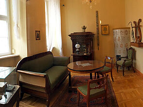 In einer Stube stehen ein historisches Sofa, zwei Sessel und ein runder Tisch. In der Ecke befindet sich ein Kachelofen. An der Wand steht ein dunkler, geöffneter Sekretär. 
