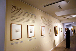 Sieben Bilder hängen an einer langen Wand und werden von einer Besucherin betrachtet.