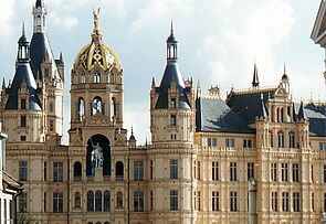 Außenaufnahme vom Schweriner Schloss. Der Ausschnitt startet im dritten Stock, erfasst das Reiterstandbild und mehrere Türme und Türmchen. Links und rechts deuten sich Fassaden historischer Gebäude aus der Schlossstraße an - so, als grenzten sie unmittelbar ans Schloss.