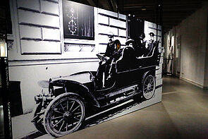 Ein XXL-Foto zeigt ein altes, offenes Auto, darin sitzen sechs Personen.