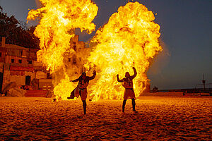 Zwei Darsteller stehen in ihren Kostümen im Sand. Hinter ihnen lodern brennende Flammen. 