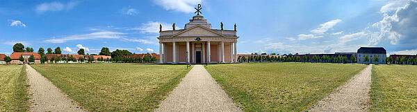 Ein Weg führt zu einem Gebäude. Das Gebäude ist die Stadtkirche. Das Gebäude hat am Eingang mehrere Säulen. Auf dem Dach stehen Figuren. Und ein Monogramm mit zwei griechischen Buchstaben.