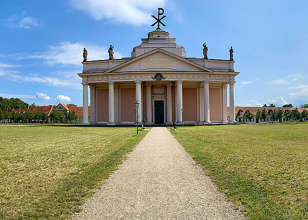 Ein Weg führt zu einem Gebäude. Das Gebäude ist die Stadtkirche. Das Gebäude hat am Eingang mehrere Säulen. Auf dem Dach stehen Figuren. Und ein Monogramm mit zwei griechischen Buchstaben.