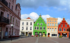 Ein Marktplatz. Vier Giebelhäuser stehen nebeneinander. Ihre Fassaden sehen weiß, grün, orange und rot aus.