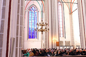 Blick in den Dom. Frauen und Männer sitzen auf Kirchenbänken.