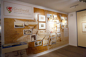 Eine Wandbild mit den Grundrissen von Greifswald. Darauf verteilt: Bilder der Ausstellung.