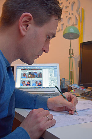 Stefan Pede sitzt an einem Tisch und zeichnet. Im Hintergrund: ein aufgestelltes Tablet mit bunten Comicheften von ihm. 