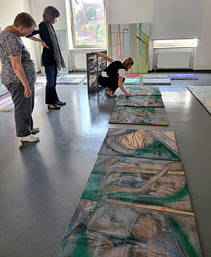 3 Frauen stehen vor einer auf dem Boden liegende Reihe an Kunstwerken. 