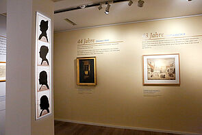 Zwei Bilder hängen an der Wand. Links an der Säule hängen Porträts von vier Frauen.