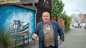 Ein Mann steht in einer Straße. Er hält ein Fischbrötchen in der Hand. Neben ihn ist eine Hauswand. Auf die Hauswand ist ein Boot gemalt. 