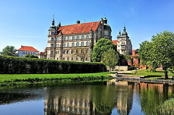 Im Vordergrund befindet sich ein Wasserlauf. Hintergrund steht ein Schloss. Rundherum blüht viel Grün.