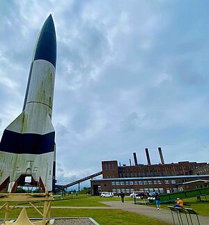 Auf einer Wiese steht ein großes Modell einer Rakete. Im Hintergrund das alte Kraftwerk.