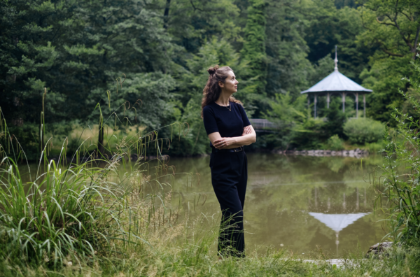 Eine Frau steht im Grünen vor einem kleinen See. Im Hintergrund ist ein Pavillon zu sehen.