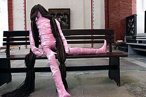 Eine Stofffigur mit langen Haaren sitzt auf einer Bank.