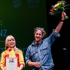 Ein Mann und eine Frau stehen auf einer Bühne. Sie sind vom Verein Freundeskreis Popkultur. Sie haben eine Urkunde in der Hand. Und Blumen. Der Mann und die Frau freuen sich.