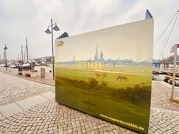 An der Promenade vom Museumshafen steht ein großes Plakat. Es zeigt die Silhouette von Greifswald, umgeben von Wiese, Bäumen und einigen Pferden. Auf dem Plakat steht auch die Internetadresse zum Jubiläum. Sie lautet www.caspardavid250.de. 
