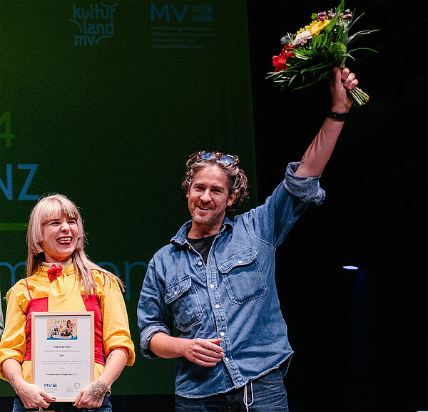 Ein Mann und eine Frau stehen auf einer Bühne. Die Frau hält eine Urkunde in der Hand. Der Mann hält einen Blumenstrauß in der Hand.