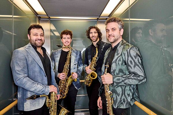 Vier Männer stehen in einem Fahrstuhl. Jeder hält ein Saxophon in der Hand.