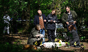 Ein Wald. Auf dem Boden liegt eine Leiche. Die Ermittler und die Rechtsmedizinerin betrachten den Tatort. 