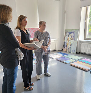 3 Frauen stehen in einem Raum und schauen nach rechts. Im Hintergrund liegen Arbeiten auf dem Boden.