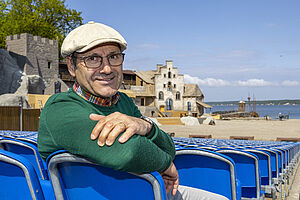 Ein Mann sitzt auf einem der Zuschauerstühle. Hinter ihm stehen Häuser aus der Kulisse. Vor den Zuschauerreihen befindet sich Sand und Wasser. 