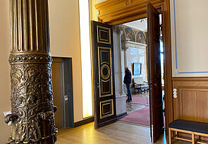 Der Eingang ins Museum führt durch eine braune Flügeltür. Gegenüber steht eine dunkle, verzierte Säule im Raum.