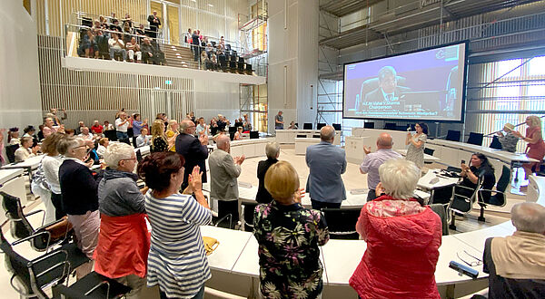 Mehrere Menschen stehen und sitzen an zu einem Rund angeordneten weißen Tischen. Sie applaudieren und zeigen fröhliche Gesichter. Im Hintergrund werden an einer Leinwand Menschen von der Live-Übertragung der Welterbeverkündung aus Indien gezeigt.