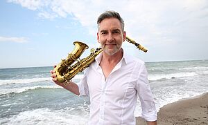 Andreas Pasternack mit seinem Saxophone über der Schulter an der Ostsee im Hintergrund.