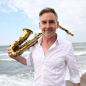 Andreas Pasternack mit seinem Saxophone über der Schulter an der Ostsee im Hintergrund.