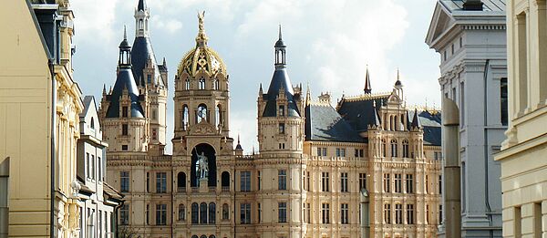 Außenaufnahme vom Schweriner Schloss. Der Ausschnitt startet im dritten Stock, erfasst das Reiterstandbild und mehrere Türme und Türmchen. Links und rechts deuten sich Fassaden historischer Gebäude aus der Schlossstraße an.